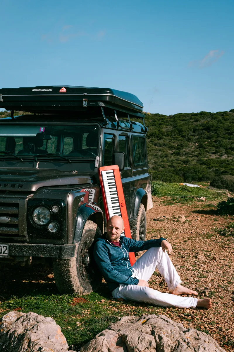 Stefan Ettinger sitzt entspannt mit einem roten Klavier an seinem Land Rover Defender in der Natur Portugals